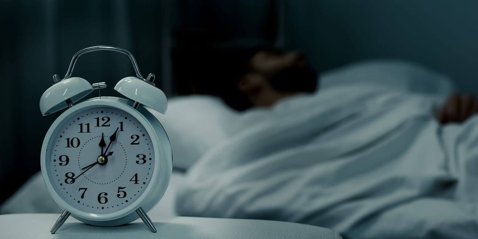 Budilka na nočni omarici v zatemnjeni spalnici, v ozadju v postelji leži oseba